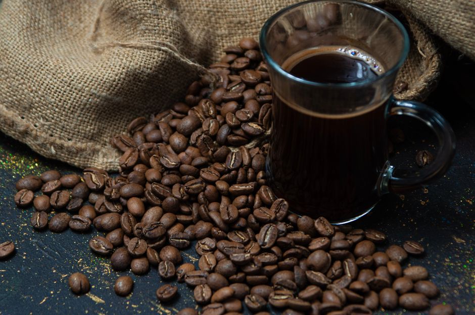 A steaming glass mug of dark coffee surrounded by roasted coffee beans and a burlap sack.